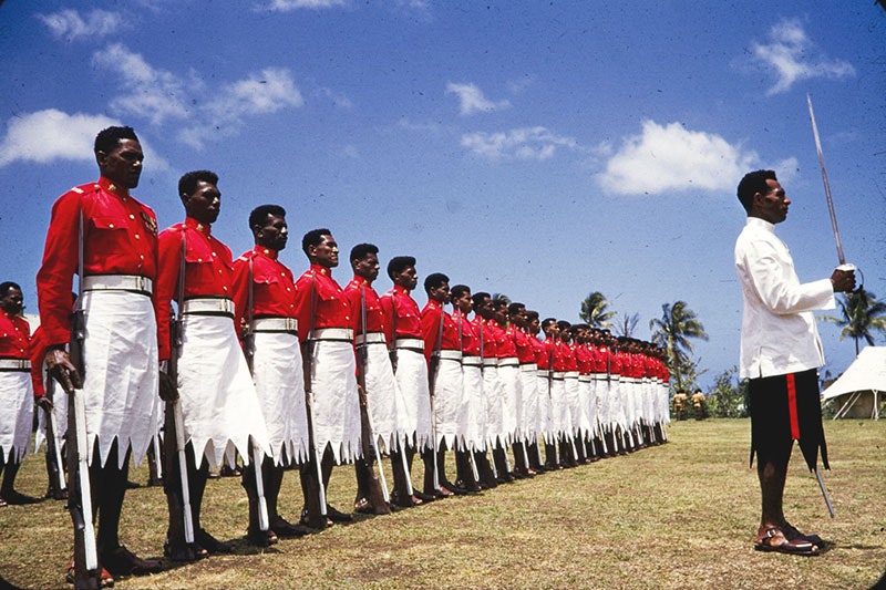 Photo of soldiers: A solitary soldier stands at the front, wearing a white coat and black sulu (kilt-like skirt) and holding a cutlass in front of him. Behind this soldier are rows of soldiers, in red shirts and white sulu, guns by their side