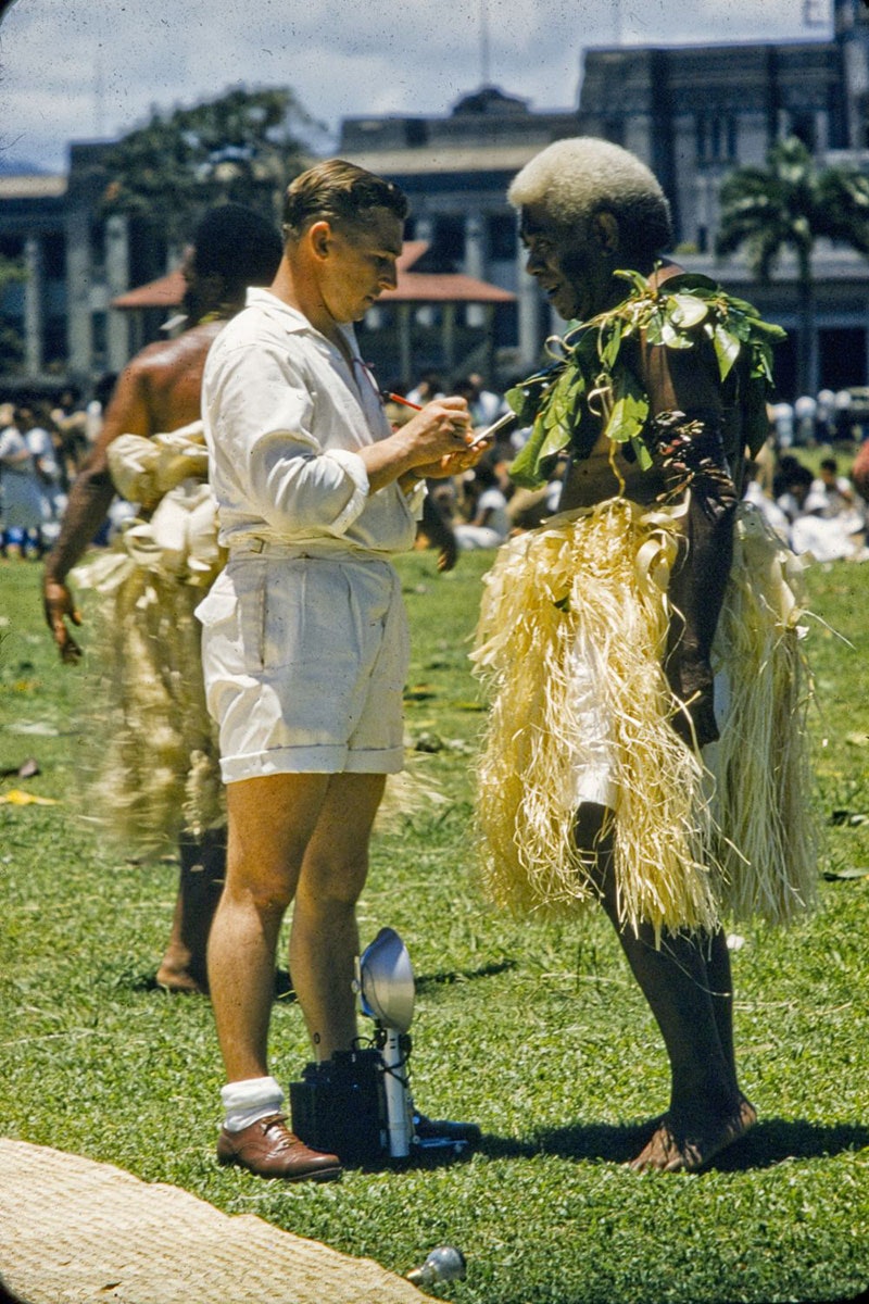 A photographer, dressed in white shirt and white shorts, takes notes as he speaks with a Fijian man