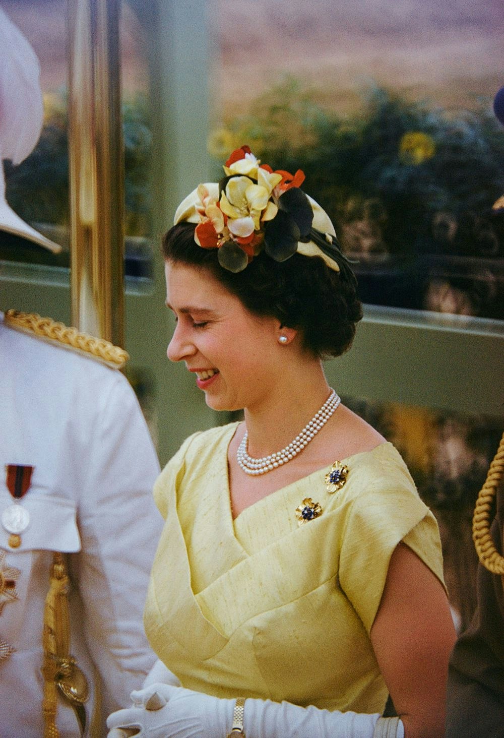 A head and shoulders of a woman in a yellow dress and floral headpiece. She is wearing white gloves and smiling at somethint
