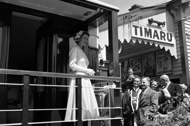 Queen Elizabeth II stands on the back of the royal train at Timaru Station. A group of people stands beside the train