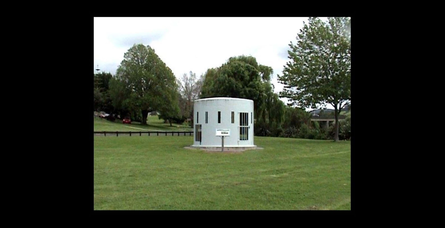 Still from a video. Pictured is a small circular building with a flat roof in a lawn. Behind it are trees, which partially obscure a building, and a road