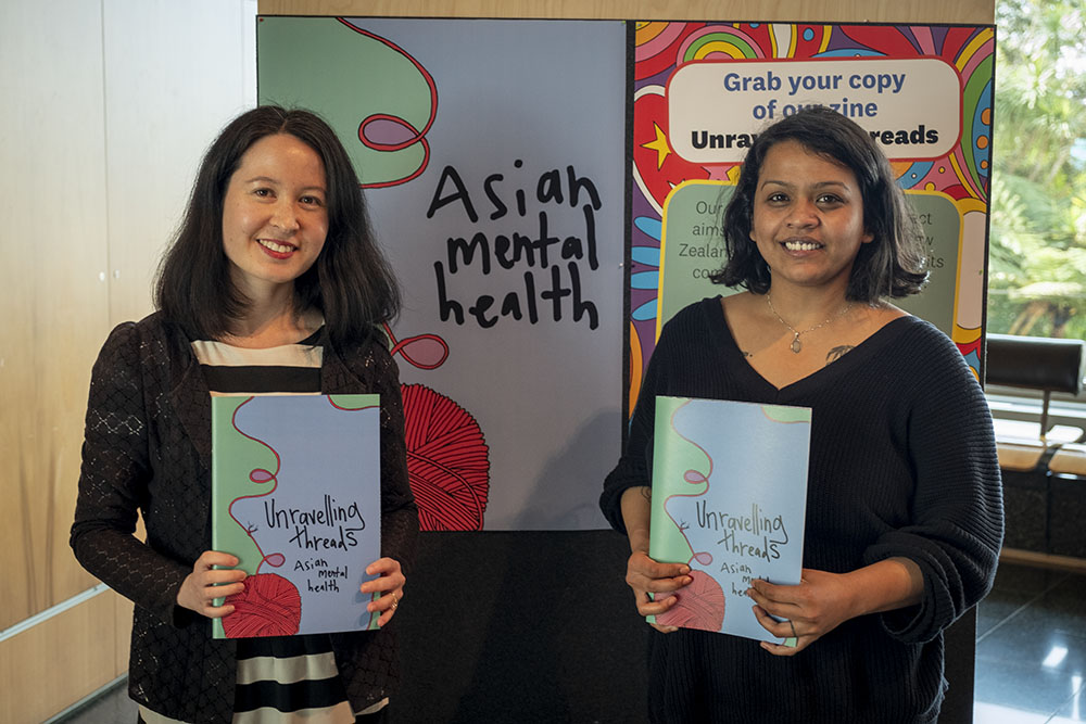 Grace and Mehwish stand in front of a sign advertising the Asian Mental Health project, holding copies of the zine
