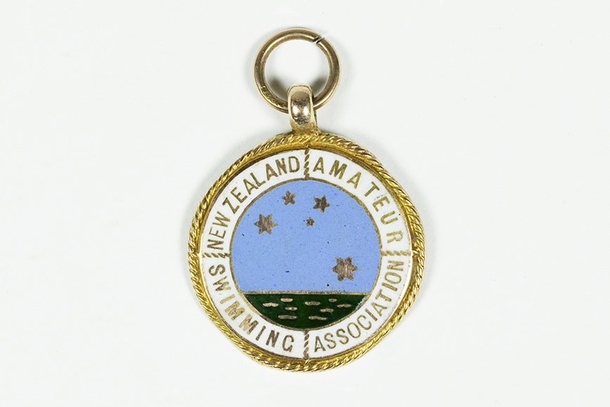 A round badge with gold edging and an image of the sea with the Southern Cross in stars above it. It has the words New Zealand Amateur Swimming Association on it