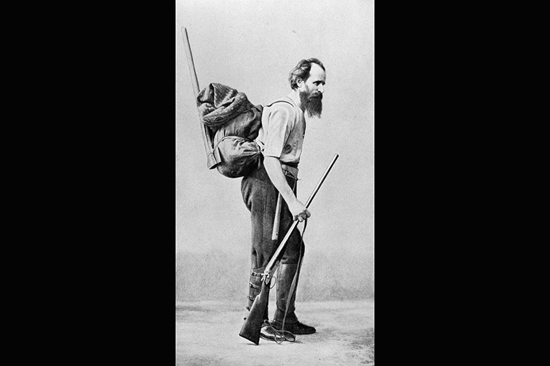 An old photo showing a bearded man carrying a pack and a rifle