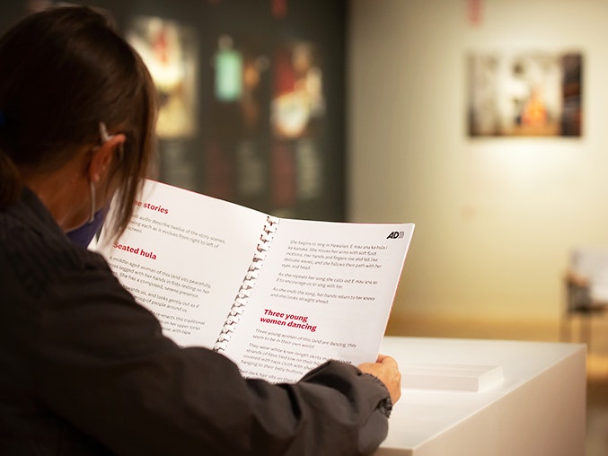 Rita Angus: New Zealand Modernist large print audio description transcript, 2022. Photo by Jo Moore. Te Papa (202422) A woman reading a large-print booklet in a gallery setting