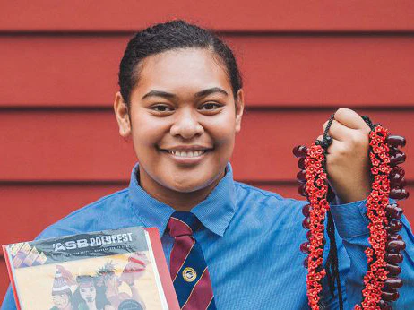 A teenage girl in a school uniform holding pandanus seed necklace and a booklet, standing in front of a dark red wooden wall