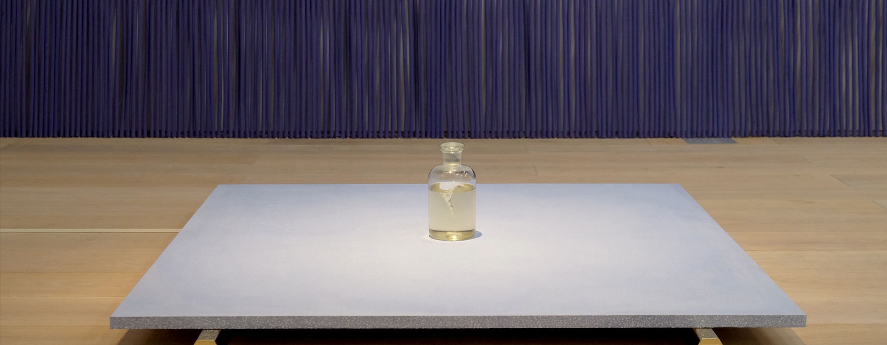A bottle of water sits on top of a light purple platform. That platform sits on top of two gold-coloured hexagonal legs. Behind, leaning against the wall is a row of over-sized incense sticks, also purple
