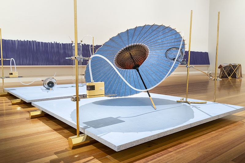 A platform sits on gold-coloured hexagonal feet. On the platform rests an iris-coloured Japanese umbrella, a camera lens and an iris flower in seperate glass jars. In the background a row of large purple incense sticks against the wall.