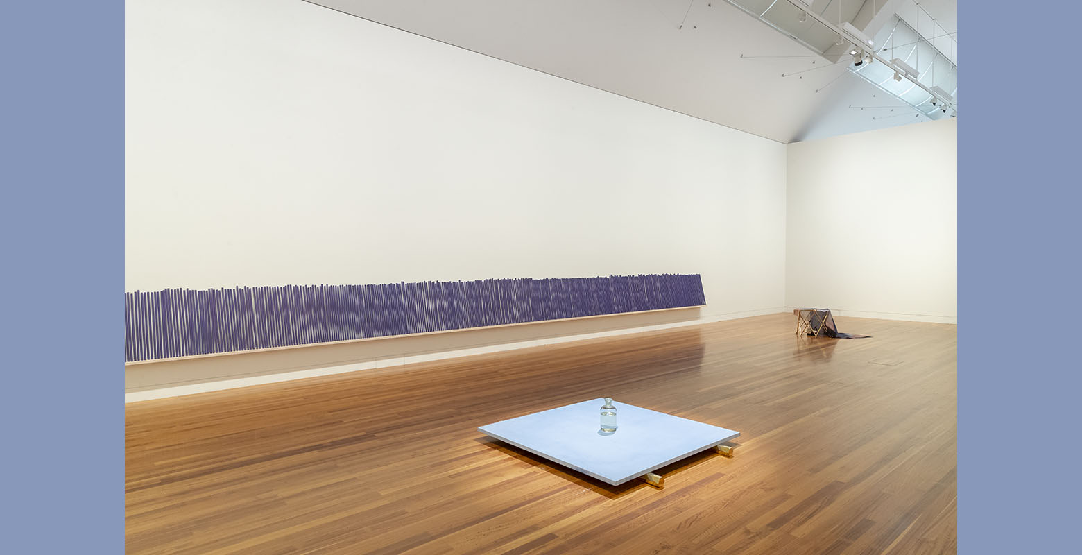 View of an expansive room with white walls and wooden floors. In the foreground, a glass jar filled with water is on a blue platform, sitting on gold hexagonal legs. Lined against the wall is a row of large purple incense sticks.