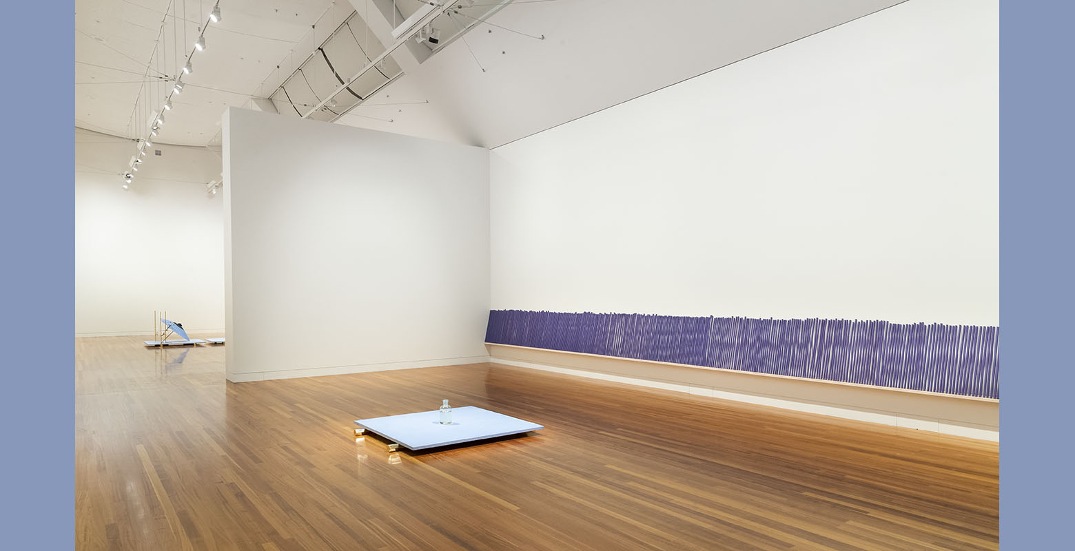 View of an expansive room with white walls and wooden floors. In the foreground, a glass jar filled with water is on a blue platform, sitting on gold hexagonal legs. Lined against the wall is a row of large purple incense sticks.