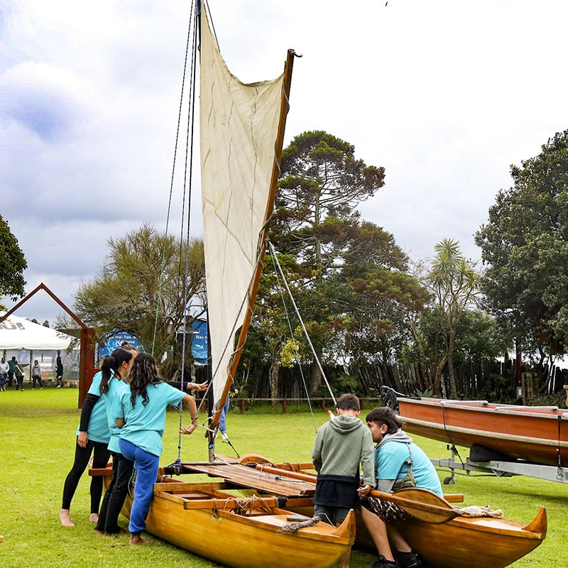 Children interact with a double-hulled waka (canoe) on a grassed area
