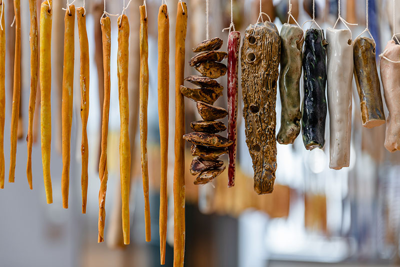 Multiple ceramic pieces hang by string, some are long and thin and dyed an orange colour; others are a variety of shapes and colours – white, dark blue, green, brown, shorter, squatter, and some have holes in them