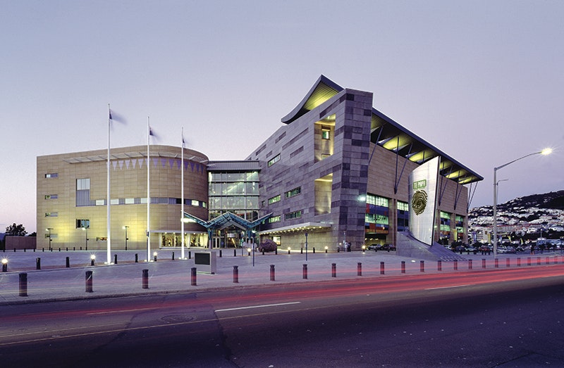 A large building that has oddly shaped walls photographed at dusk. There are three flags outside the entrance.