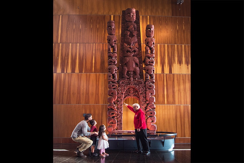 A man in a red top is pointing at a very tall wooden carving. He is describing something to the man crouched next to two small children.