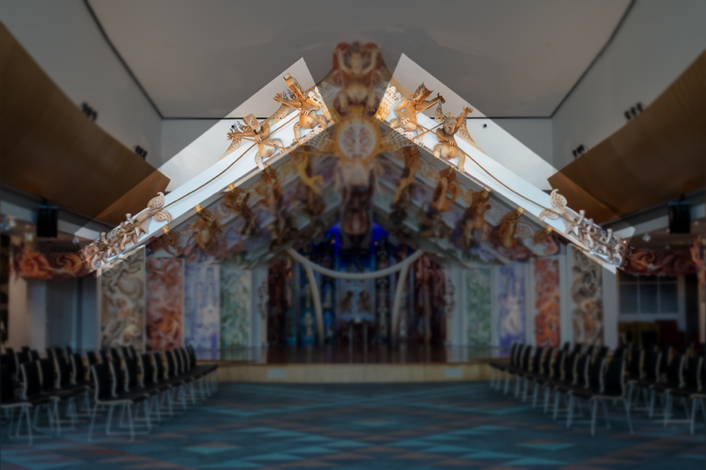 A multicoloured stage with an open-wall house at the back with carvings made of MDF on the sides and rafters. There are chairs either side of the stage set out so they face each other.  One section is highlighted the rest of the image is darkened.