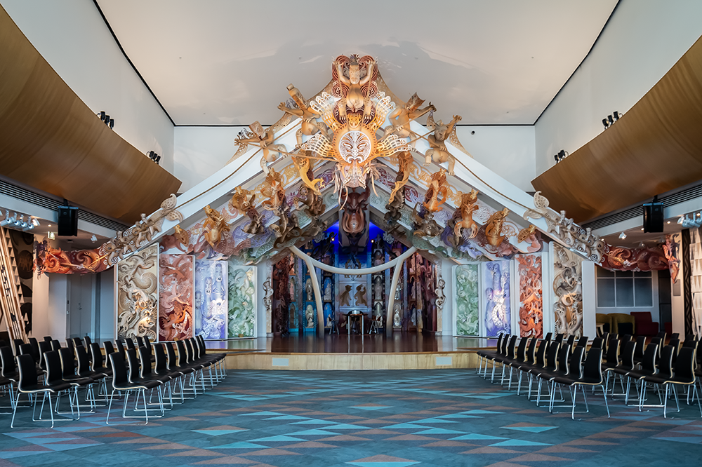 A multicoloured stage with an open-wall house at the back with carvings made of MDF on the sides and rafters. There are chairs either side of the stage set out so they face each other.