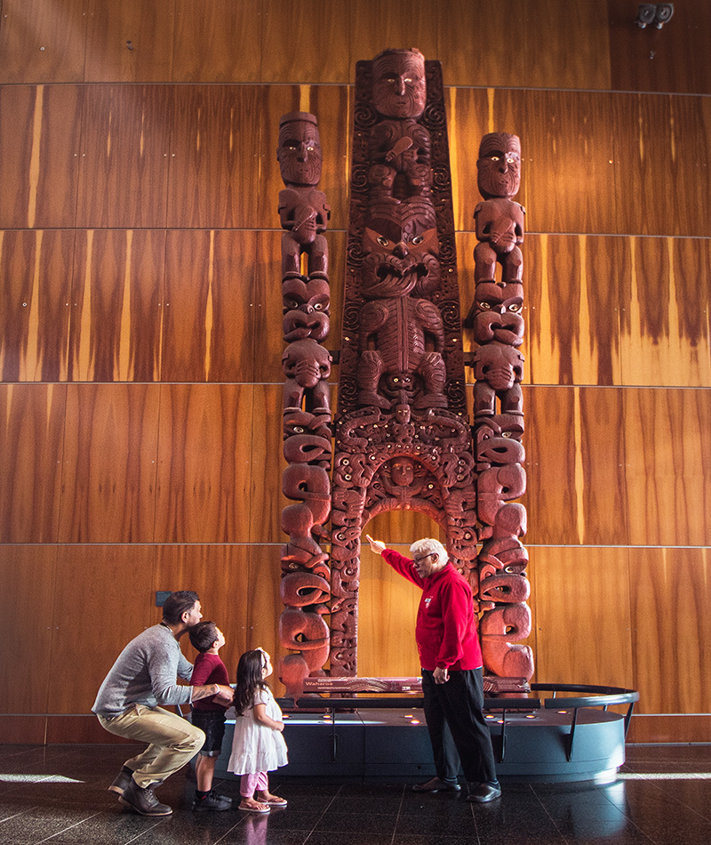 A man in a red top is pointing at a very tall wooden carving. He is describing something to the man crouched next to two small children.