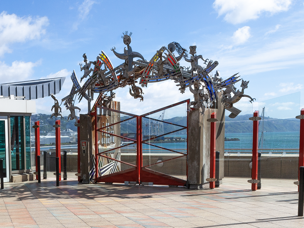 An ornamental archway over a red outdoor gate. There is a glass fence either side of it and part of a building with windows on the left.