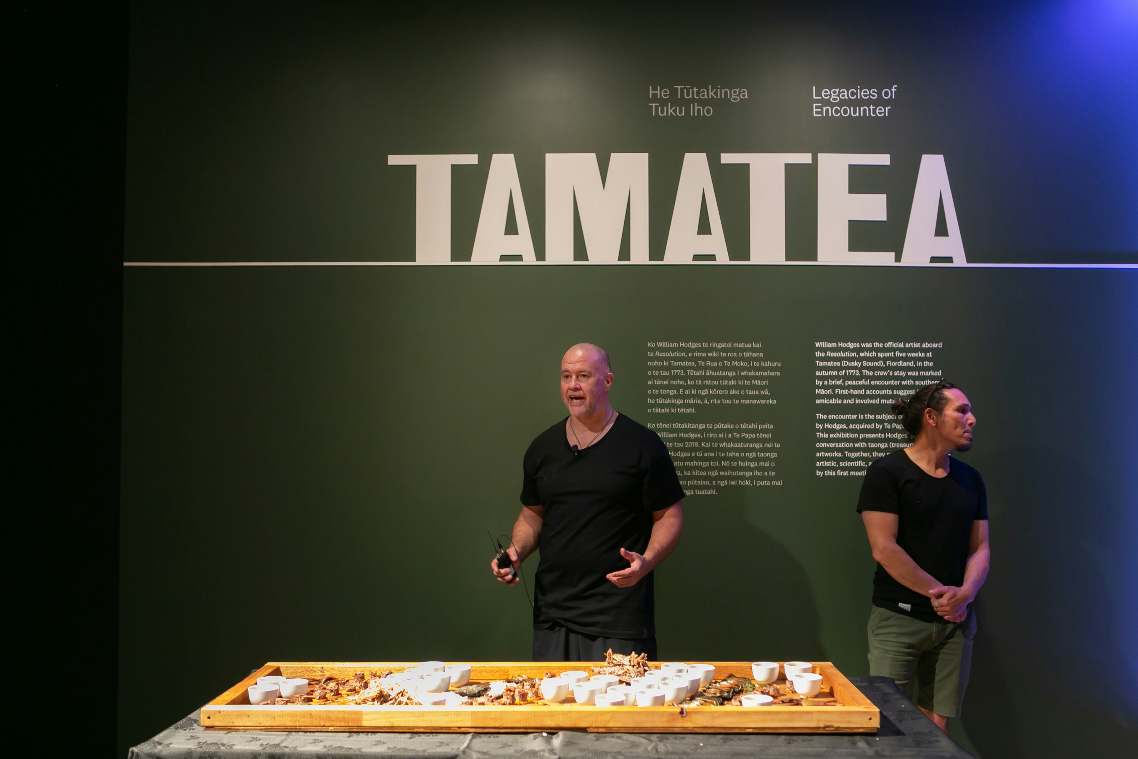 Two me stand against a green wall with the word “Tamatea” written on it, talking to a crowd behind the camera. In front of them is a table with an array of foods on it