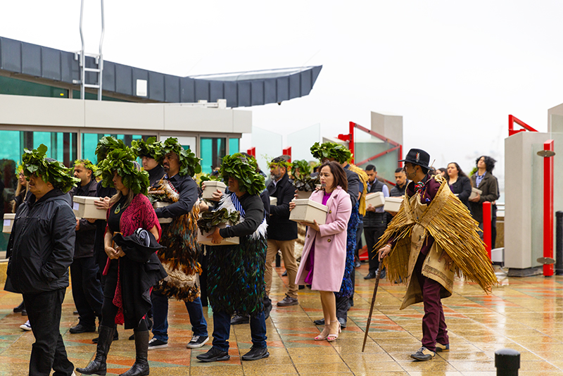 A lot of people, some carrying boxes are walking into a building. One man is wearing a top hat and woven cloak.