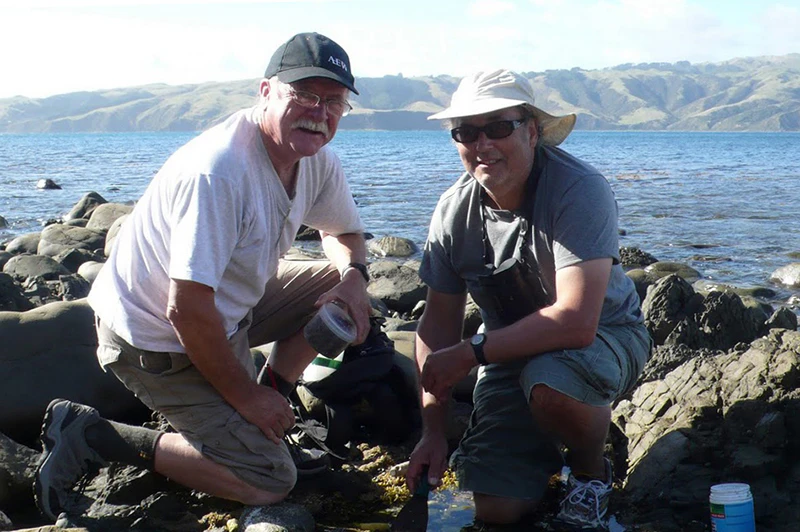 Two men are crouched on coastal rocks with jars of specimens in front of them.