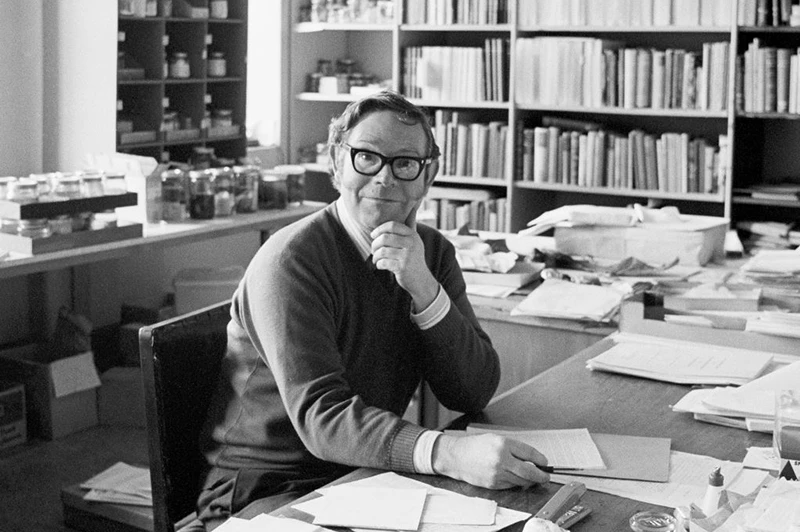 A black and white photo of a man with black rimmed glasses siting at a desk surrounded by a lot of papers.