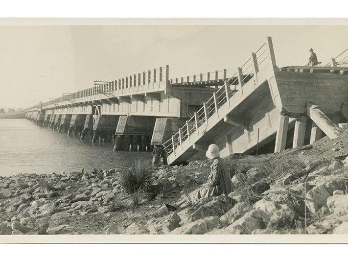 1918 road and rail bridge over the Ahuriri Estuary, Westshore, Napier, circa 1931, Hawke's Bay, maker unknown. Gift of Mrs J Paterson, date unknown. Te Papa (O.005491) A woman sits on the side of the river looking at a bridge that has collapsed after an earthquake.