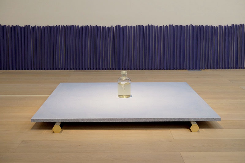 A bottle of water sits on top of a light purple platform. That platform sits on top of two gold-coloured hexagonal legs. Behind, leaning against the wall is a row of over-sized incense sticks, also purple