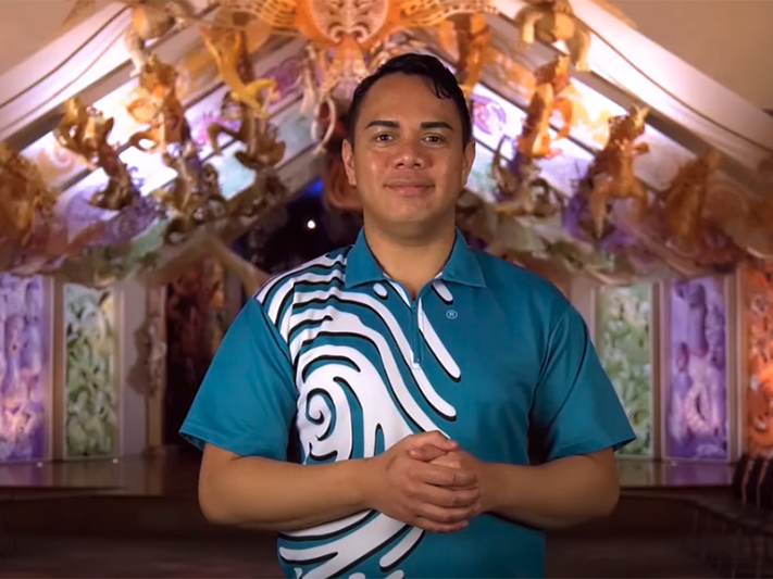 NZSL Introduction to Rongomaraeroa, 2021. Te Papa (168115) Sponsored by the New Zealand Sign Language Fund A man standing in front of a carved building with his hands clasped in front of him.