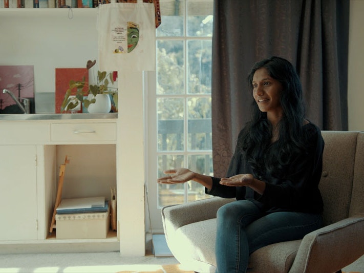 Still from ‘Healing doesn’t just happen in one mode’: reconsidering what ‘therapy’ looks like, 2023. Photo by Julie Zhu Video still of a woman sitting on a chair talking to somebody off-camera. Behind her is a window with curtain partially drawn, and a bench with lots of books on it