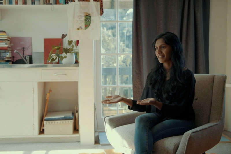 Video still of a woman sitting on a chair talking to somebody off-camera. Behind her is a window with curtain partially drawn, and a bench with lots of books on it