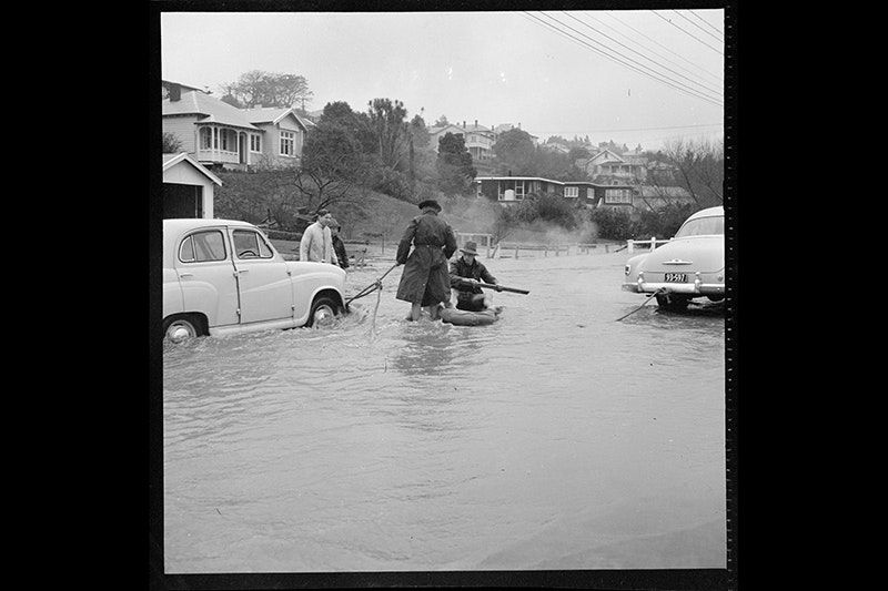 Two people are tying a rope to a car in a flooded street. There are two other people looking on.