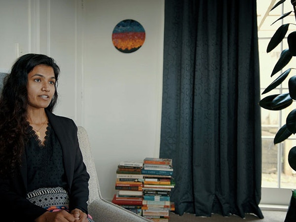 Sehar Moughal, 2022. Photo by Julie Zhu A woman sits on a light grey chair, talking to somewhere out of the frame on the right. Behind her sits two piles of books, and a painting on the wall