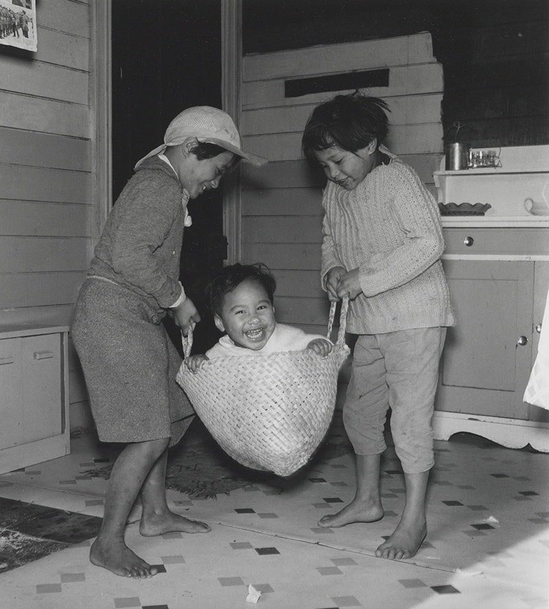 Photograph of two children swinging a baby in a flax kete or basket.