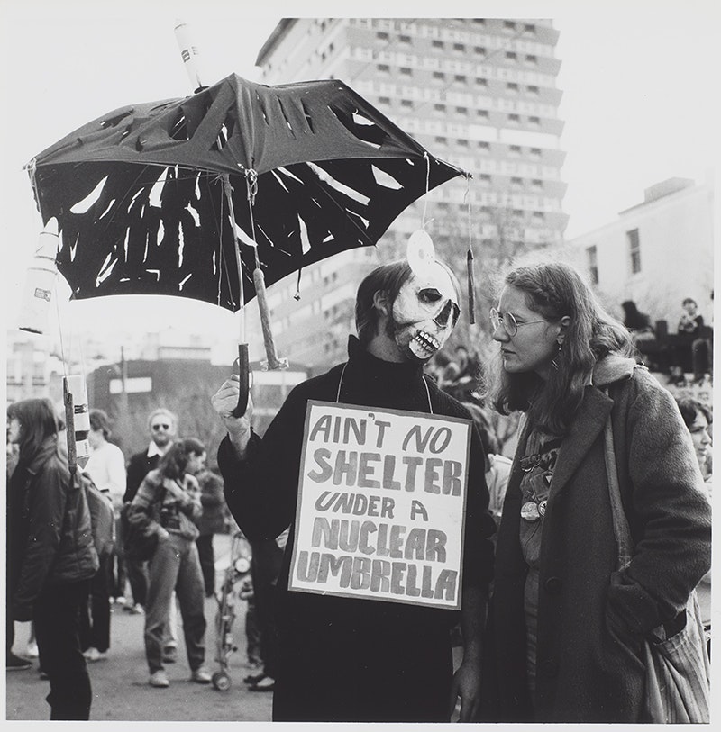 Two people talking while walking in a march. One person has a face mask on and is holding a sign that says "Ain't no shelter under a nuclear umbrella".