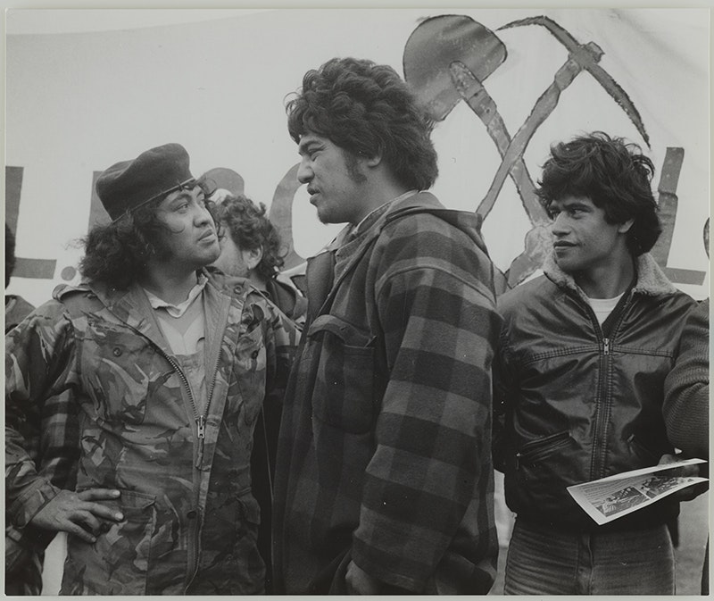 Three men standing outdoors. Two are looking at each other and the third is looking on.