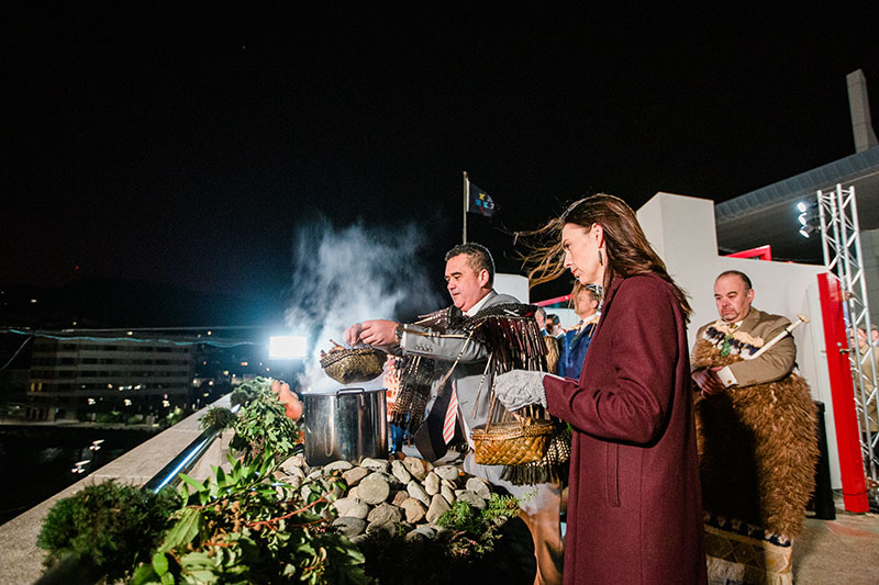 A man and a woman stand outside. It’s early morning and the sky is still dark. The man lifts food out of the pot, releasing steam.