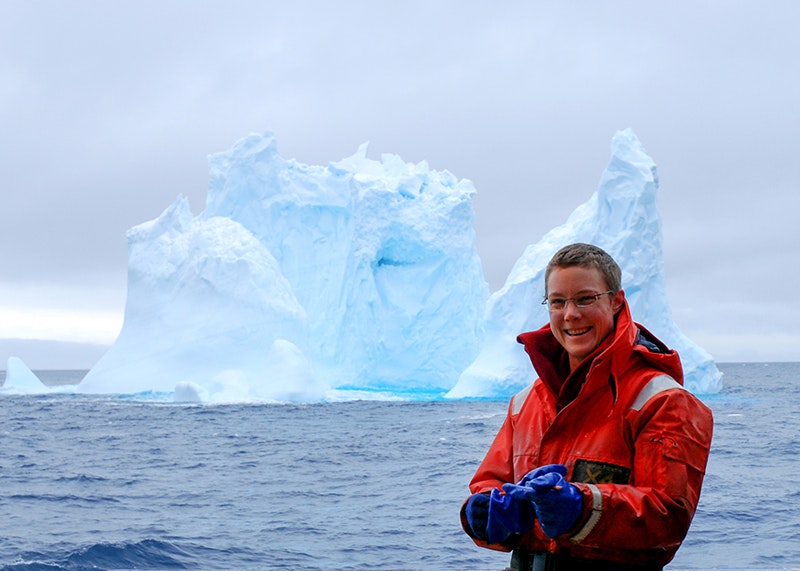 A woman stands in front of a large iceberg. There is sea between her and the iceberg.