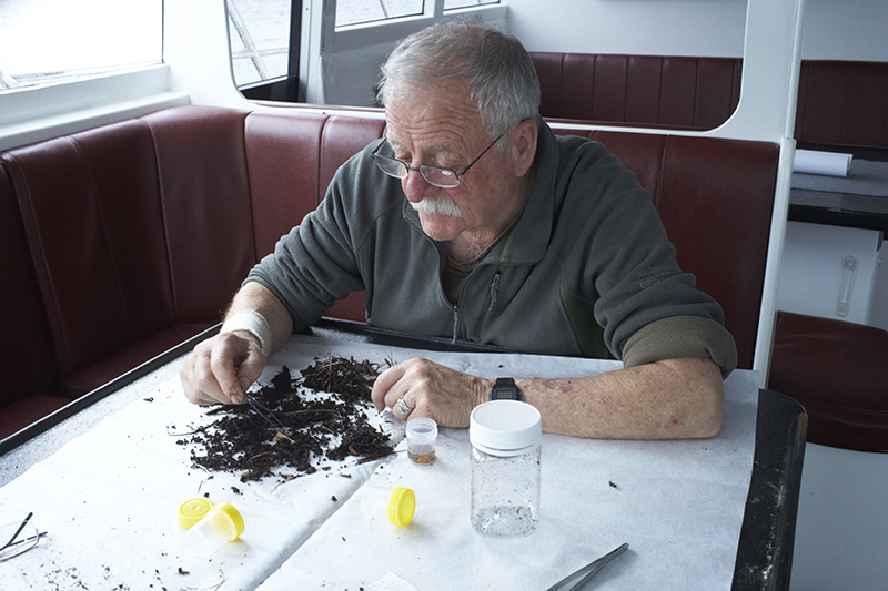 A man is sitting at a table on a boat sifting through a selection of dirt with tweezers.