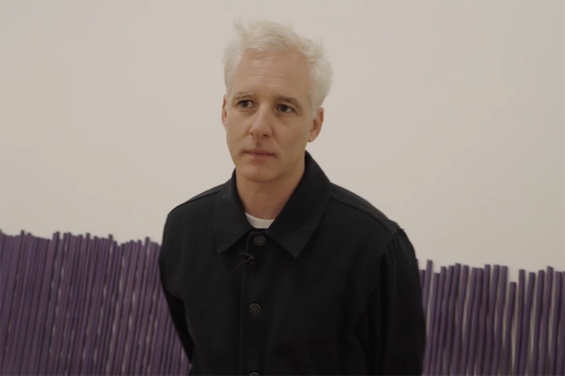 A man with white hair is standing in a room that has large purple incense sticks lying against the wall.
