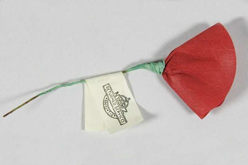 A red paper flower representing a poppy with a green stalk made of wire. There is a tag on the stalk with the words Returned Services Association and their emblem.