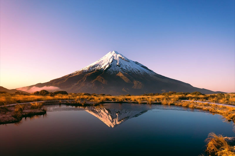 View of conical Mt Taranaki, with a body of water in front of it reflecting the mountain