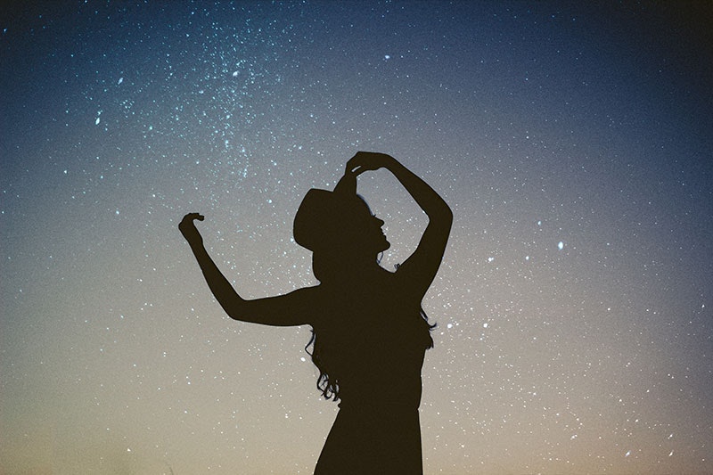 Silhouette of a woman holding her brimmed hat on her head, with the stars in the sky behind her