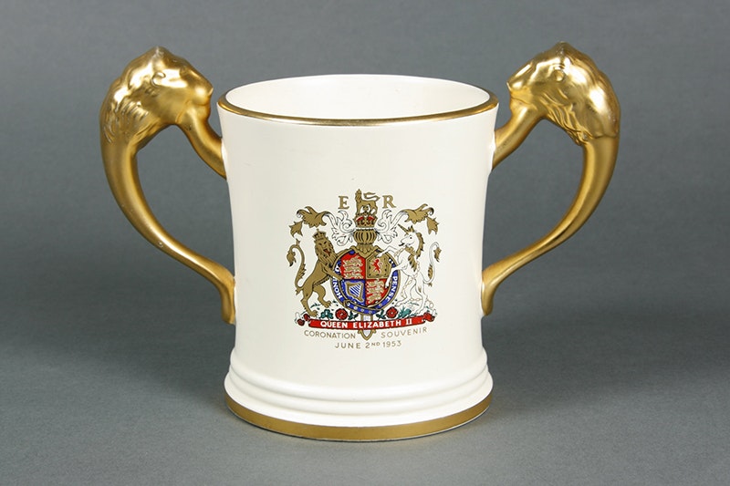 A white cup with gold handles on either side and a coat of arms in the middle.