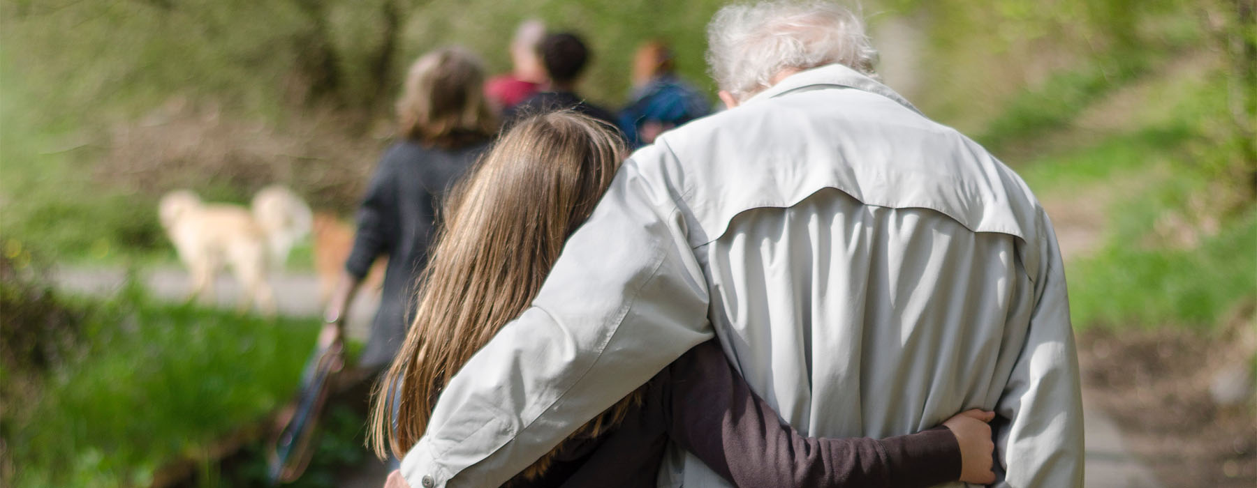 A young girl and an older man – possibly her grandfather – walk arm in arm along a path surrounded by trees
