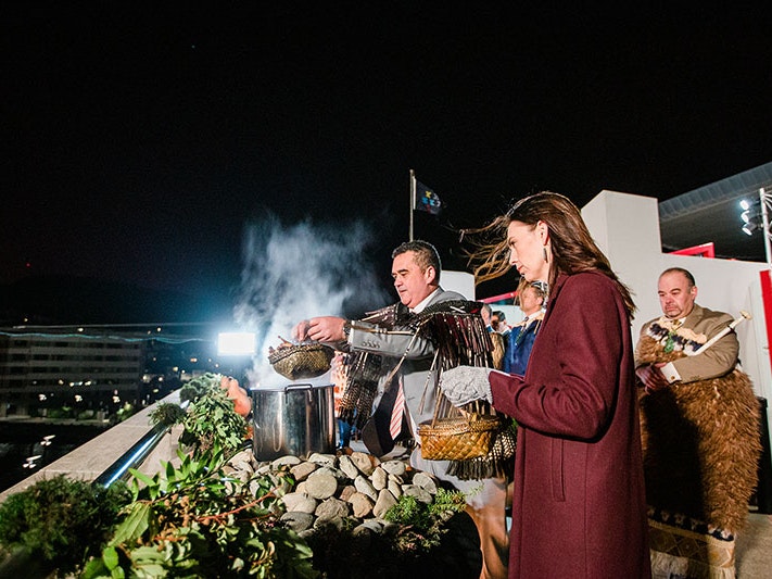 Rangi Mātāmua lifts kai (food) from the cooking pot as it releasing steam into the sky, with then-Prime Minister Jacinda Ardern looking on, during the whāngai i te hautapu during Matariki, 24 Jun 2022. Photo by Erica Sinclair. Te Papa (210553) Rangi Mātāmua lifts the lid from the food pot to release steam into the sky, with then-Prime Minister Jacinda Ardern looking on
