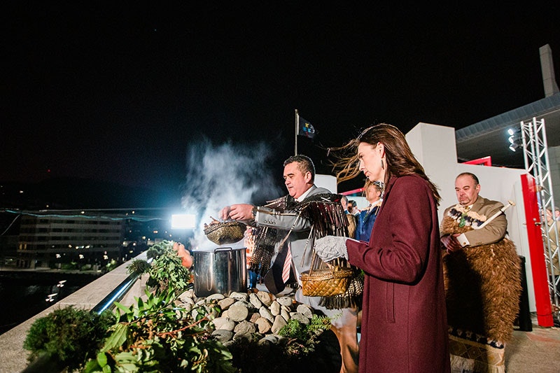 Rangi Mātāmua lifts the lid from the food pot to release steam into the sky, with then-Prime Minister Jacinda Ardern looking on