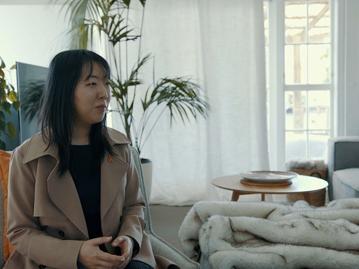 Rebekah Jaung, 2022. Photo by Julie Zhu. Te Papa Woman wearing a beige trench coat sits on a couch in a light-drenched room. Behind her is a TV set, to her right is a large couch with a blanket draped over it, and glass doors behind it