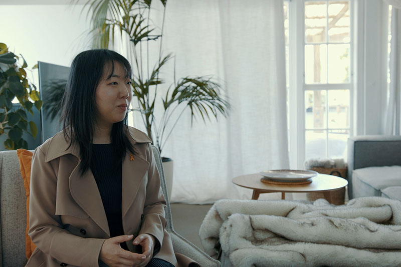 Woman wearing a beige trench coat sits on a couch in a light-drenched room. Behind her is a TV set, to her right is a large couch with a blanket draped over it, and glass doors behind it