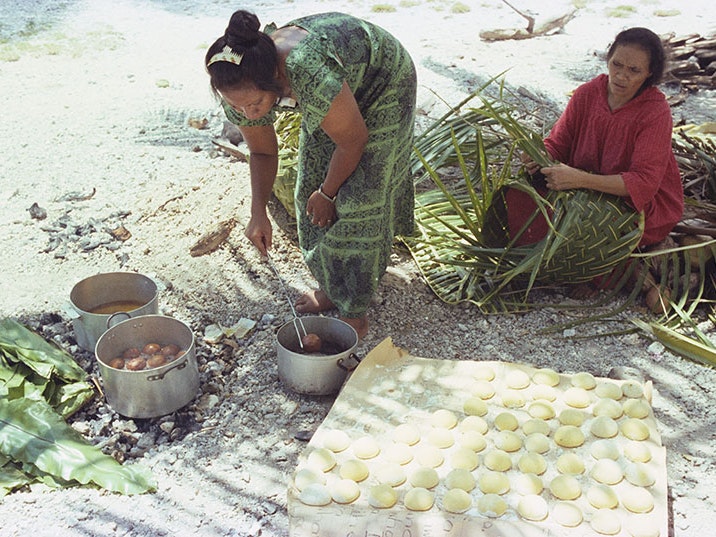 Glenn Jowitt, Woman cooking panikeke, 1981, Tokelau, colour negative. Gift of Glenn Jowitt Estate, 2015. Te Papa (E.007346) A woman prepares bread to fry in a pot, while another woman weaves with flax. They are sitting on a beach